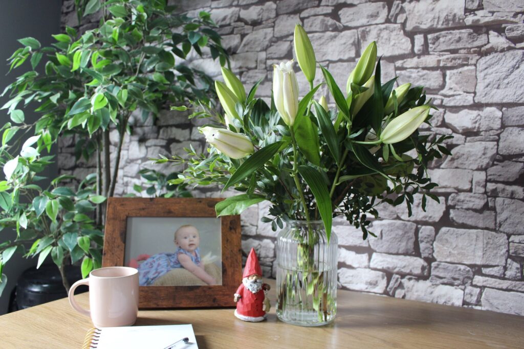 A bouquet of lilies in a glass vase on a wooden desk with photo frame and notepad. 5 benefits of flower shopping locally