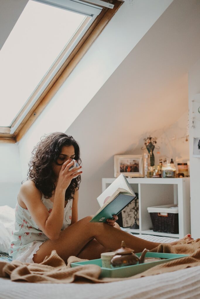 Woman sat on bed drinking a cup of tea and reading a book. This is to go with an article discussing different ways to refresh your self care regime.