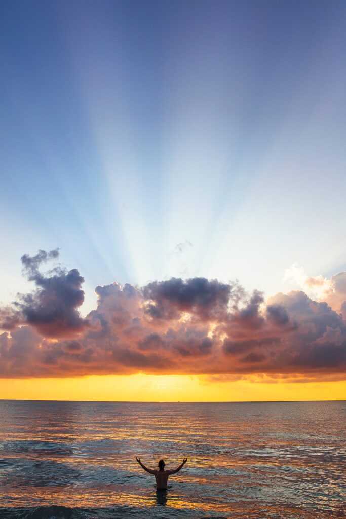 Man standing in ocean raising hands in faith towards the sunrise.