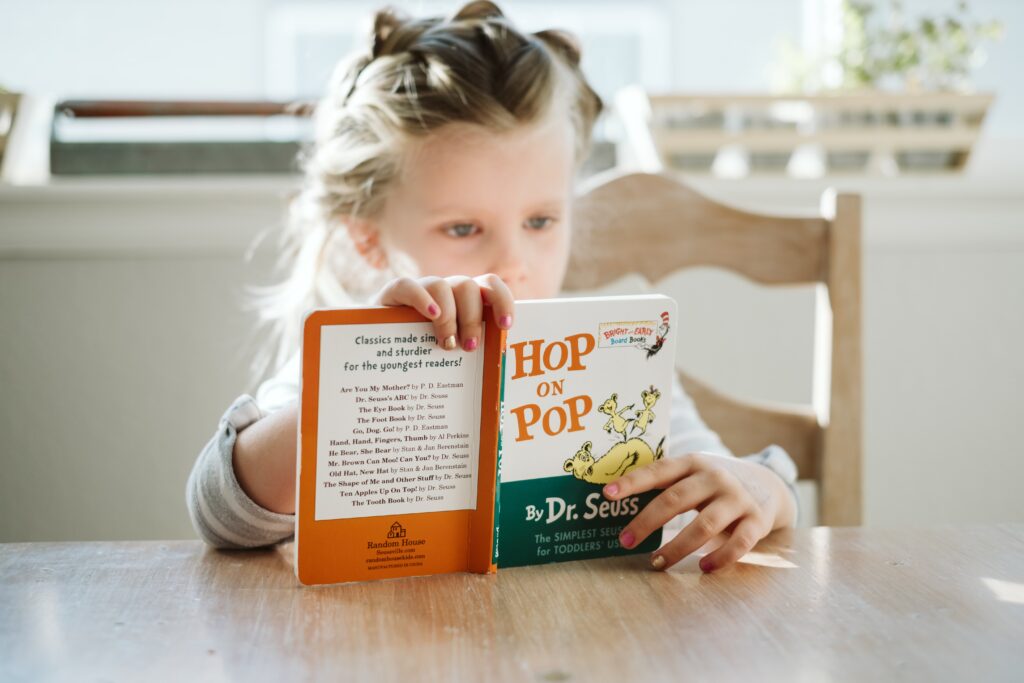 Little girl sat at dining table reading. This image is to go with an article giving tips for teaching your child to read.