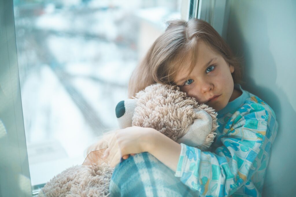 Little girl sat in window cuddling teddy bear.