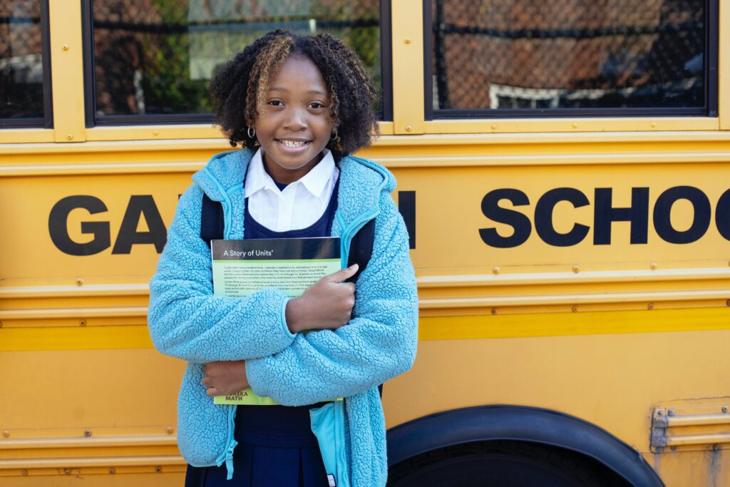 Young girl holding textbook stood in front of a yellow school bus. This image refers to an article discussing how to help your child prepare for the school bus.