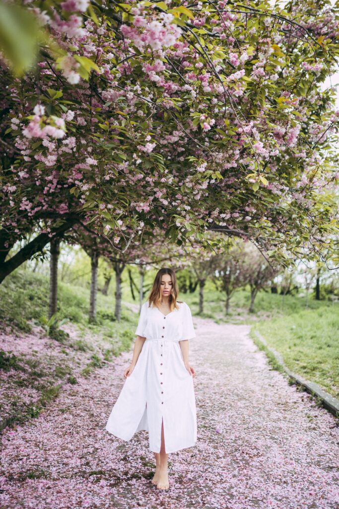 Blonde woman walking through blossom filled park wearing a white spring capsule wardrobe dress.