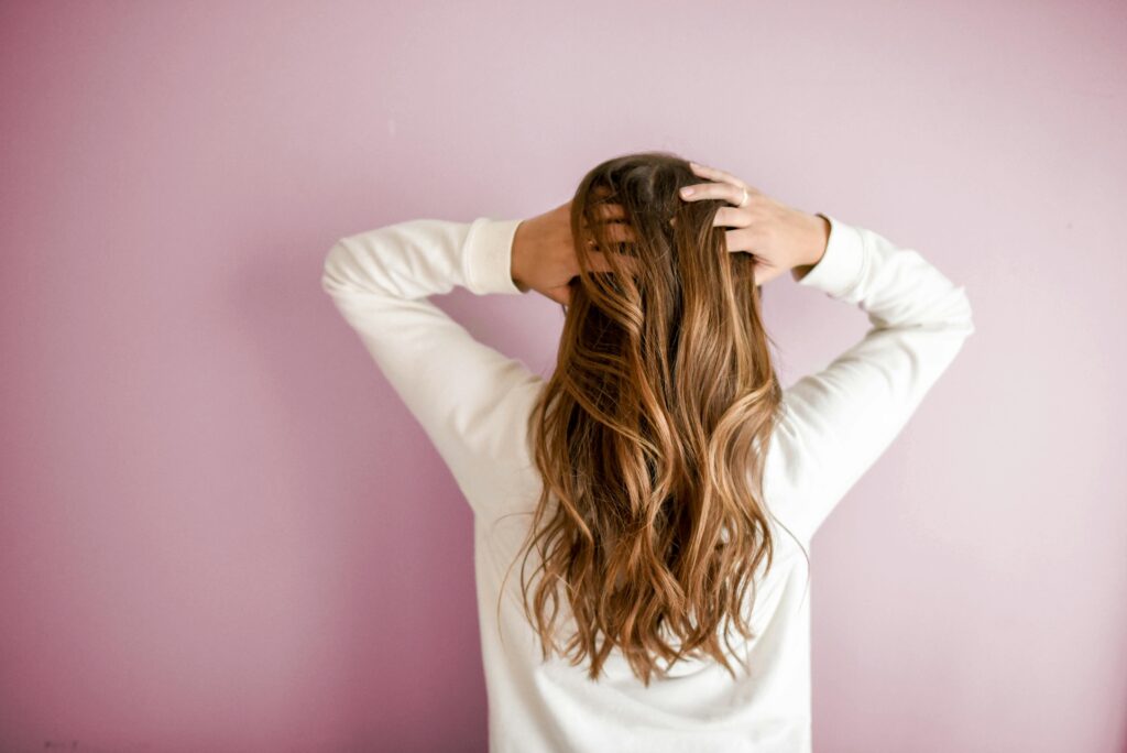 Pink background with a woman standing looking away from camera running hands through her hair. This is to go with an article discussing postpartum hair truths.