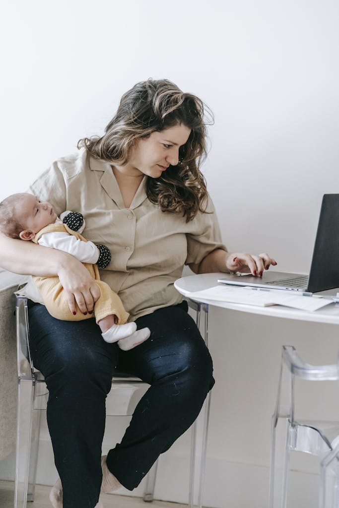 Concentrated mom sitting on chair with cute baby and browsing laptop while working remotely on maternity leave
