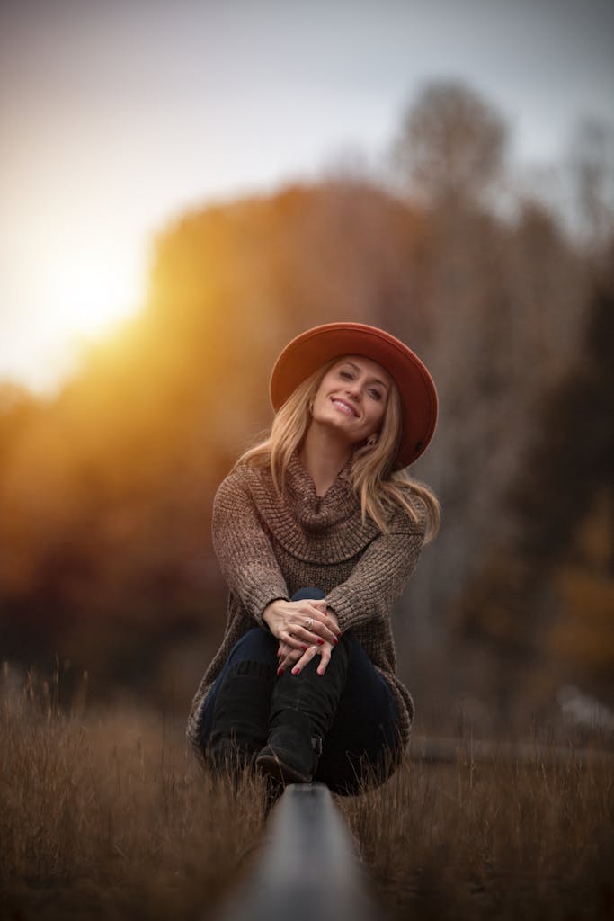 A cheerful woman in autumn attire sitting outdoors, bathed in warm sunset light.