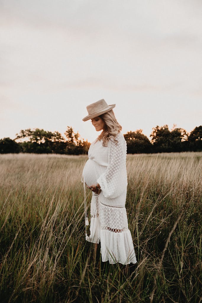 Stylish pregnant woman in a white dress and hat standing in a grassy field during sunset.